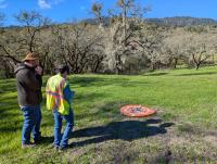 Two people prepare to fly a drone at the cemetery. One person is on walkie-talkie and the other has the controls for the drone, which sits on an orange launchpad