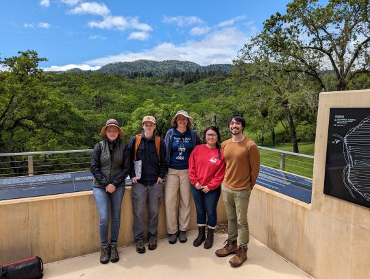 Dr. Alexis Boutin (tall White woman) stands with four students (two on each side of her) on concrete memorial platform at Home Cemetery. Behind them are cemetery, trees, and hills.