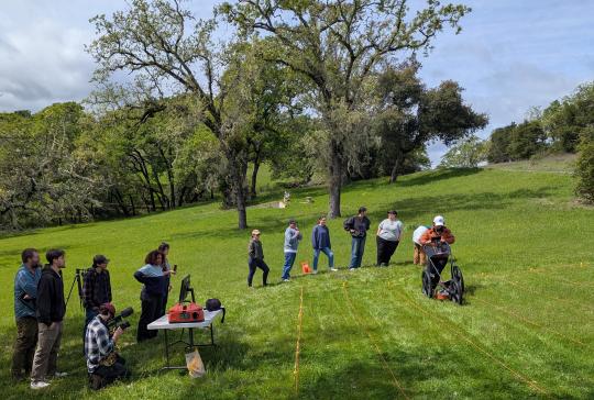 Students conducting geophysical survey with ground-penetrating radar at the Sonoma Developmental Center cemetery. One student pushes the GPR unit across the grass while ten others look on. 