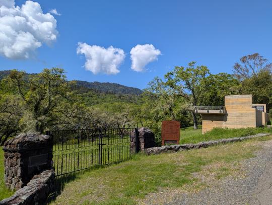 Iron gates and stone columns and low wall in front of Home Cemetery. Concrete memorial platform is visible to the right, overlooking the cemetery. The cemetery appears to be an open field covered in green vegetation, with oak trees and the Sonoma Mountains visible in the background.