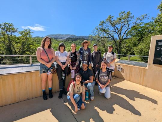 Dr. Boutin and nine Sonoma State students smile at the camera while visiting the SDC Home Cemetery memorial platform. Trees and hills are visible in the distance behind them. 